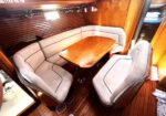 Dinette area inside a wooden boat cabin: curved light-gray cushioned benches around a polished wooden table.