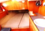 Wood-paneled boat cabin with a V-berth bed and two cushions, small sink to the right, a wall fan, and a fire extinguisher mounted on the left.