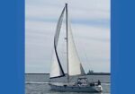 Sailboat with white sails on calm water, heading near a distant breakwater under a blue sky.