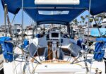 Rear cockpit of a white sailing yacht with a wooden deck, steering wheel, and blue canopy, docked in a sunny marina with other boats in the background.