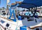 Sailboat docked at a marina with a blue canopy/bimini, cockpit seating, and coiled ropes, with other boats in the background.