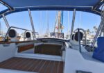 View from the cockpit of a sailboat in a marina, with wooden deck, white rails, and blue canopy; masts and other boats in the background.