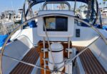 Sailboat cockpit view: steering wheel, chartplotter screen at the helm, wooden pedestal, teak deck, with moored boats in a marina behind.