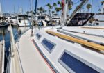 Sailboat deck at a marina with other boats, palm trees, and a bright blue sky; hull windows and a wooden railing run along the side.