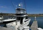 White motor yacht moored at a wooden dock in a calm harbor, with blue water and a distant shore on a sunny day.