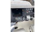 View of a boat dashboard at the helm: a large screen display, several gauges, radio and controls, throttle levers, and a person’s feet in white pants and black shoes visible nearby.