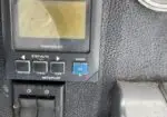 Close-up of an airplane autopilot panel with a small display screen and a blue Mode button on a dark textured cockpit surface.