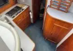 Narrow boat galley with wooden cabinetry, a stainless sink on the left, a two-burner stove, a fire extinguisher on the wall, and a curved wooden cabinet at right; blue carpeted floor.
