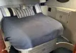 Grey-blue bed with striped pillows in a compact boat cabin, storage drawers beneath the bed edge.