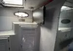 White boat cabin corner with vented cabinet, wall-mounted TV, circular porthole window, and skylight above.