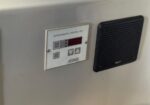 Environmental control unit panel with a digital display and buttons mounted on a gray wall, beside a black speaker grill.