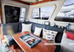 Cozy boat cabin with a blue upholstered L-shaped sofa, wood table, cushions, and a blue tray of magazines; windows overlook a marina.