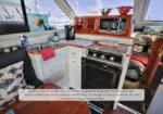 Compact yacht galley with white cabinets, wood trim, oven, microwave, sink, and a seating area nearby. Marina visible through windows in the background.