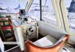 Lower helm cockpit of a boat with steering wheel, gauges, and navigation screen, marina visible through windows in the background.