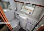 Compact marine bathroom with a sink, mirror, storage cabinets, and a full-size head (toilet) in a small gray/white cabin with wood trim fixtures.