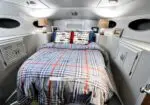Owner's cabin forward aboard a yacht: a bed with plaid blue-red bedding, two lamps, portholes, and built-in cabinets.