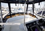 Cockpit of a motorboat with white seating and control panel; marina with docked yachts visible through the cabin windows.