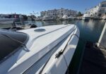 White motorboat docked at a marina, tied to a wooden pier, with calm water, other boats, and coastal apartment buildings in the background.