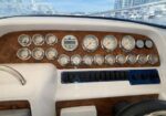 Boat cockpit dashboard with many round analog gauges on a wood veneer panel and a row of switches or toggles beneath it, steering wheel partially visible at right edge.