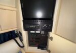 Cabinet with an open door showing a small screen monitor above a row of switches and controls on a boat’s electronics panel, plus a mounted phone handset nearby.