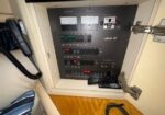 Electrical control panel with meters and switches inside an open white cabinet; a remote control rests on a ledge and a coiled black cord lies on the wooden surface.