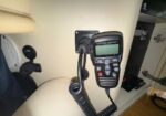 Handheld radio transceiver mounted on an aircraft cabin wall with a coiled cord and keypad display for cockpit communications.