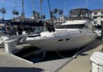 White motor yacht moored at a marina, with other boats, palm trees, and low-rise buildings in the background.