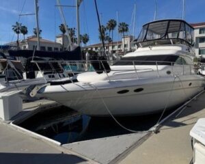 White motor yacht moored at a marina, with other boats, palm trees, and low-rise buildings in the background.
