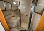 Small boat bathroom with a curved sink, mirror cabinet, enclosed shower with a grab bar, porthole window, wooden door frame, and a teal bath mat