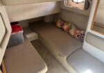 Compact boat cabin with beige seating arranged as a bed, floral cushions, a plastic storage bin, a small drawer, and a porthole window.