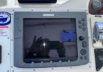 Marine navigation console with a central square display and surrounding control buttons; photographer's hands are reflected on the screen.