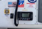 VHF marine radio mounted on a white panel, with a coiled mic cord and multiple stickers (US flag, BoatUS, celebration sticker) on the surface of the console.