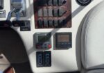 Close-up of a vehicle cockpit control panel with many switches and two small digital displays next to a beige seat cushion.