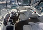 Captain's helm on a white yacht: steering wheel, gauge panel, and captain chair with additional seating in a sunlit cockpit.