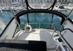View from the cockpit of a small yacht at a marina, with a lifebuoy on the deck and a curved glass enclosure around the helm area.