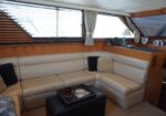 Beige L-shaped leather sofa in a boat cabin with wooden paneling, windows with blinds, and a black ottoman with magazines.