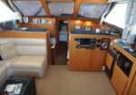 Cozy yacht salon with beige leather sofa, wood cabinets, and a galley area; navigation panel and controls on the right, with large windows and curtains above.