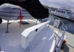 Deck of a white sailboat in a marina; emergency liferaft canister mounted on the deck with red straps, surrounded by stainless rails and nearby docked boats.