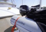 Inflatable dinghy labeled Caribe attached to a larger sailboat, with a Suzuki four-stroke outboard motor and a black cover, at a marina.