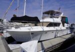White motor yacht moored at a marina with a black cover over the forward deck and blue sky above.