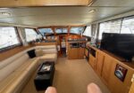 Lounge area of a wooden yacht: beige leather sectional, black ottoman coffee table, flat-screen TV on the right, built-in cabinets, and large windows with curtains; helm area visible in the back.