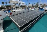 Solar panels installed on the deck of a sailboat docked in a marina, with masts and palm trees in the background.