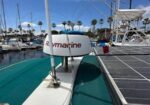 View from a sailboat deck at a marina, with a Raymarine navigation unit, fishing rods, and surrounding boats under a blue sky with palm trees in the distance.