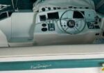 Boat cockpit helm with steering wheel and navigation dash, gauges and controls under a protective cover; teal exterior with a lower storage shelf containing cups and cleaning supplies.