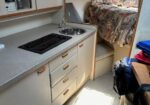 Compact boat galley with a stove, sink, and cream cabinets along a counter, leading to a sleeping berth with a floral bedspread in the background.