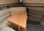 Cozy boat cabin with U-shaped beige cushioned seating and a rectangular wooden table, small window above, storage shelves along the wall.