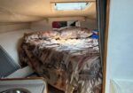 Cozy boat cabin with a bed under a window hatch, patterned duvet, and a small kitchenette sink to the left.