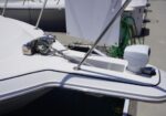 Close-up of a white boat bow at a marina with an anchor chain, windlass, and deck hardware; green hoses and dock equipment visible in the background.