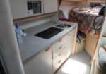 Compact caravan kitchenette with a two-burner stove, sink, and microwave above white cabinets; bed area visible in the back.