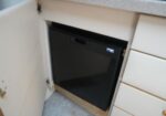 Compact black dishwasher installed under a kitchen counter with an open cabinet door nearby.